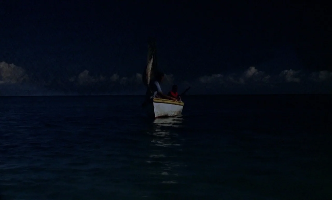 Dr. No (1962) movie still: wide — A small wooden sailboat floats on the open ocean under a dark night sky. Two individuals sit inside the small boat…