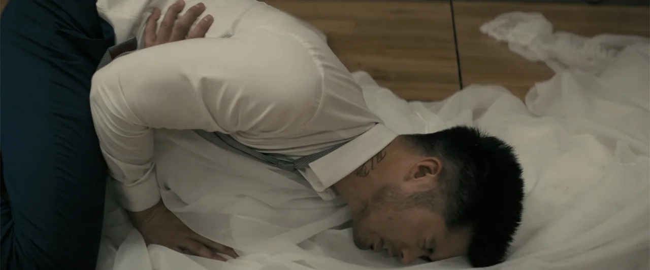 Perfect Addiction (2023) movie still: medium — A man lies prone on a bed covered with white flowing textile. He rests face down on the white fabric…