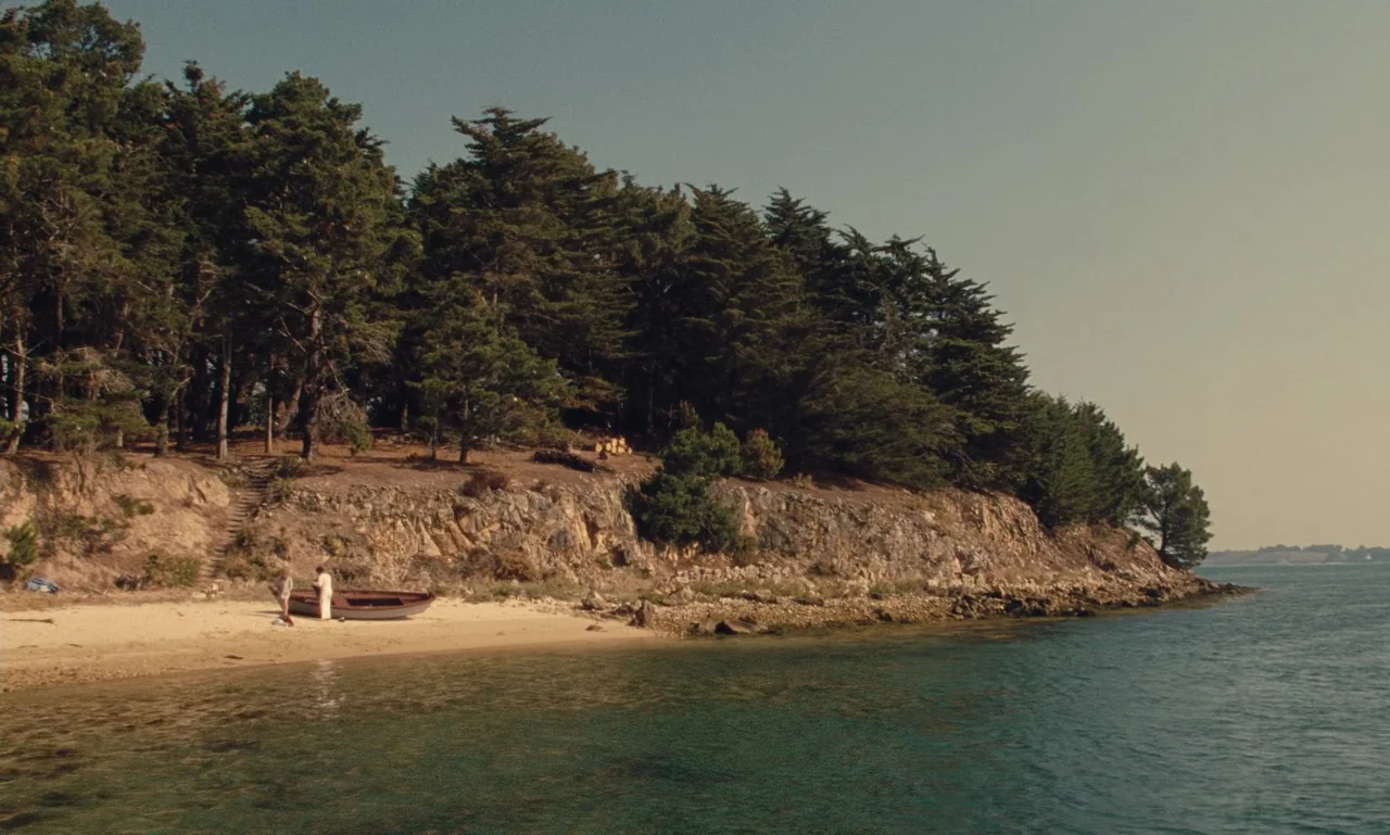 A Tale of Winter (1992) movie still: wide — A quiet, rocky coastline meets a thin sandy beach with a dense pine forest. Two figures stand beside a…