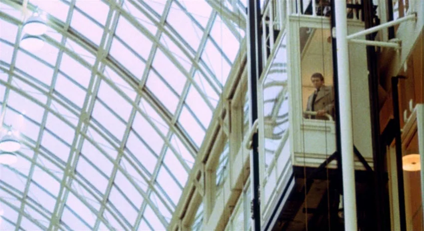 The Silent Partner (1978) movie still: wide — A modern commercial atrium features a prominent glass roof and an exposed cable elevator. A man…