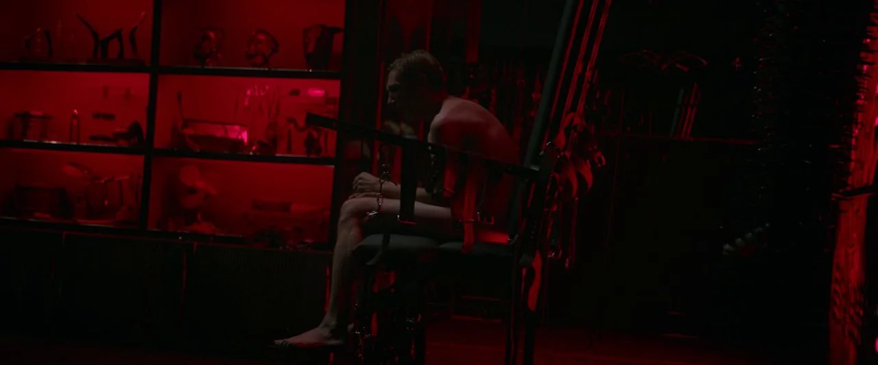 Dogs Don't Wear Pants (2019) movie still: medium — A dark, ominous room features a man strapped into a chair with shelves behind. The man rests in…