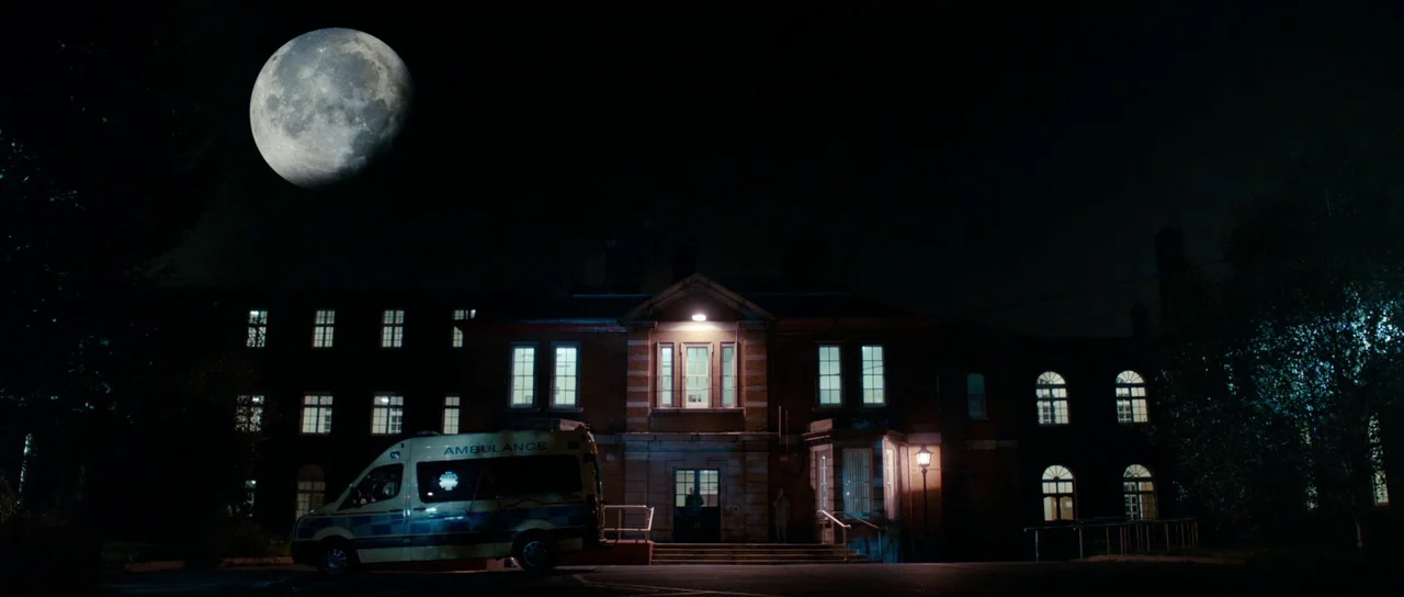 Calvary (2014) movie still: wide — A large multi-story Victorian-style hospital building stands dark under a massive full moon. An ambulance is…