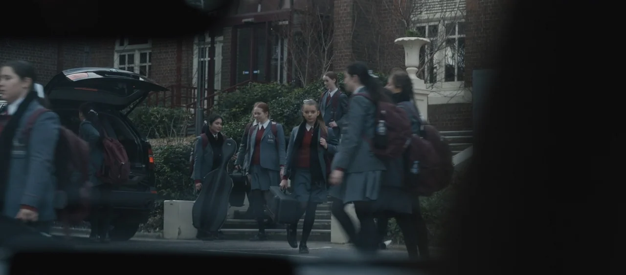 Force of Nature: The Dry 2 (2024) movie still: wide — A group of teenage students in school uniforms walk from a parked car. The brick building has…