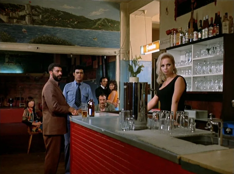 Ali: Fear Eats The Soul (1974) movie still: wide — A bar interior features a red slatted counter and a mural of a landscape. A blonde bartender…