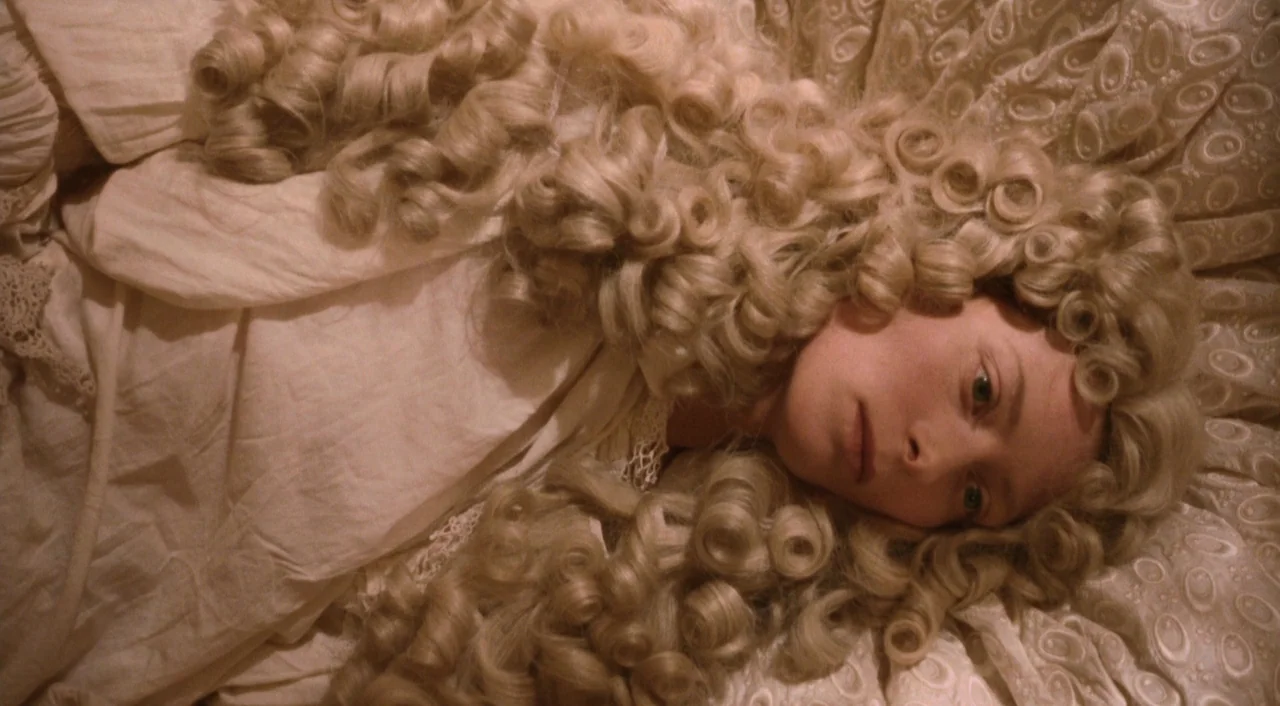 Orlando (1992) movie still: closeup — A person lies on a white patterned fabric surface while wearing a large wig. The subject rests their head…