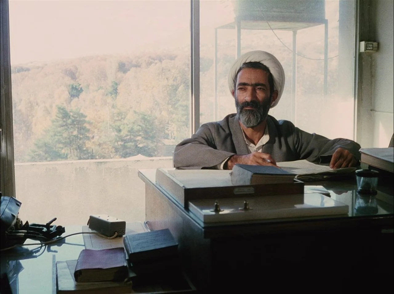 Close-Up (1990) movie still: medium — A workspace with a desk positioned in front of a large, bright window. A man in a turban sits at the desk…