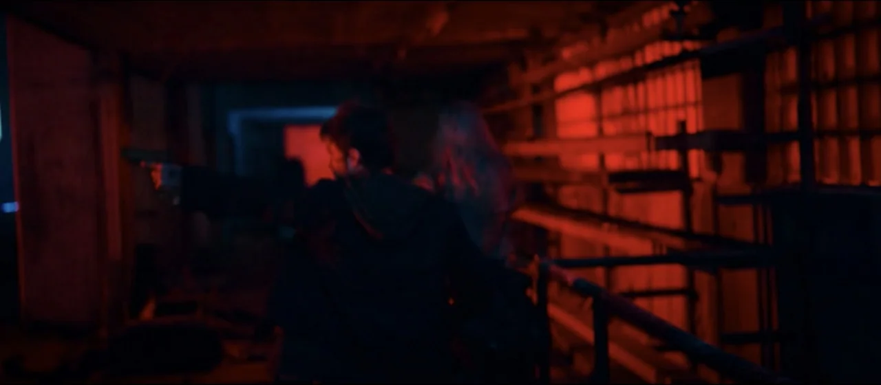 Guns Akimbo (2020) movie still: medium — An industrial hallway features metallic railings and exposed pipes under intense red illumination. A man…