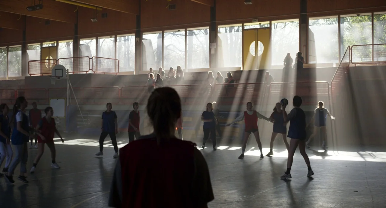 Zombi Child (2019) movie still: wide — A spacious gymnasium features high windows with strong rays of light cutting through dust. A group of…