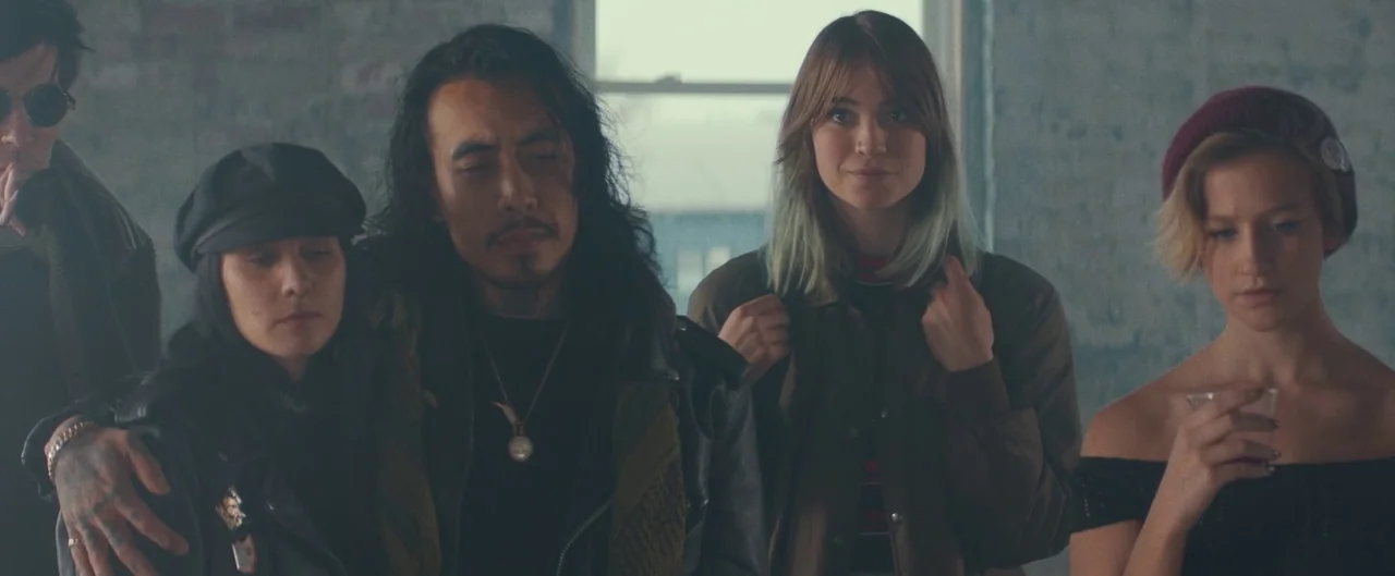 Poser (2021) movie still: medium — A spacious loft interior with grey brick walls and a large, bright window. Four young adults stand together in a…