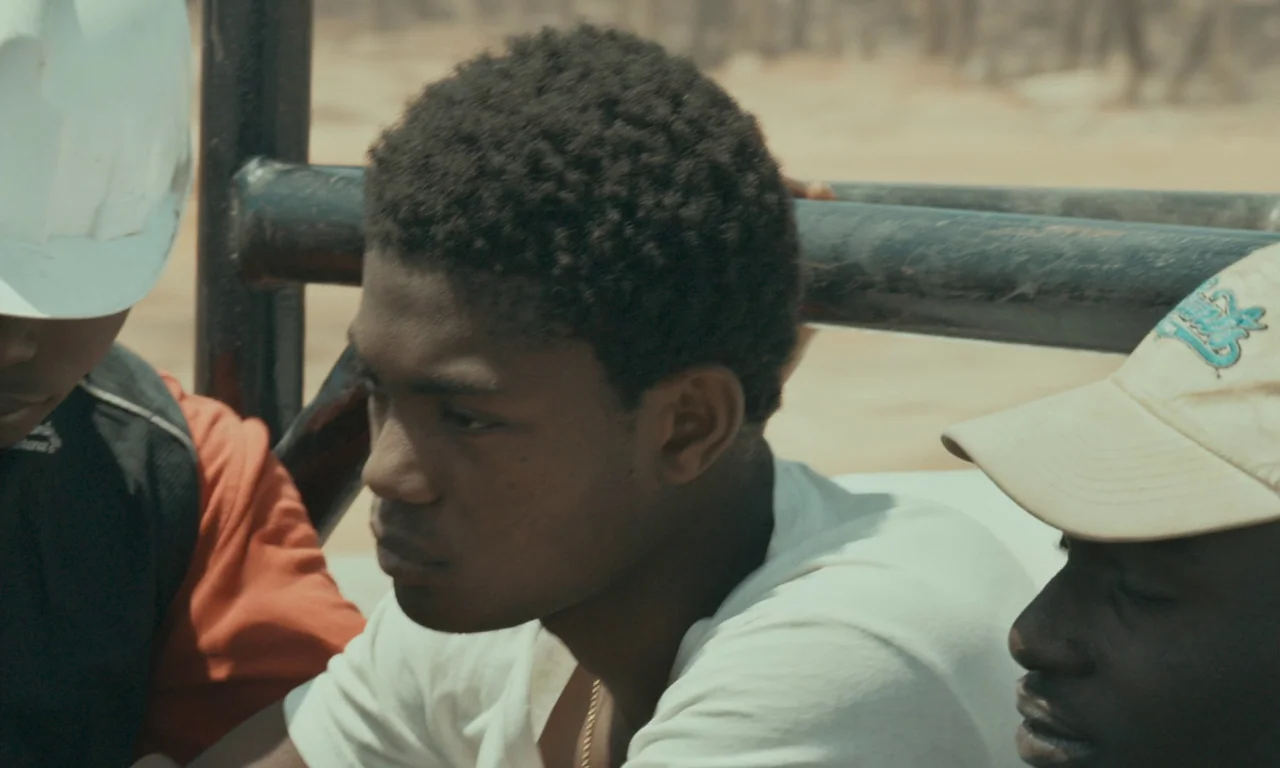 Atlantics (2019) movie still: medium — A group of young men sit together inside the back of an open vehicle. Dry, light-colored landscape visible in…