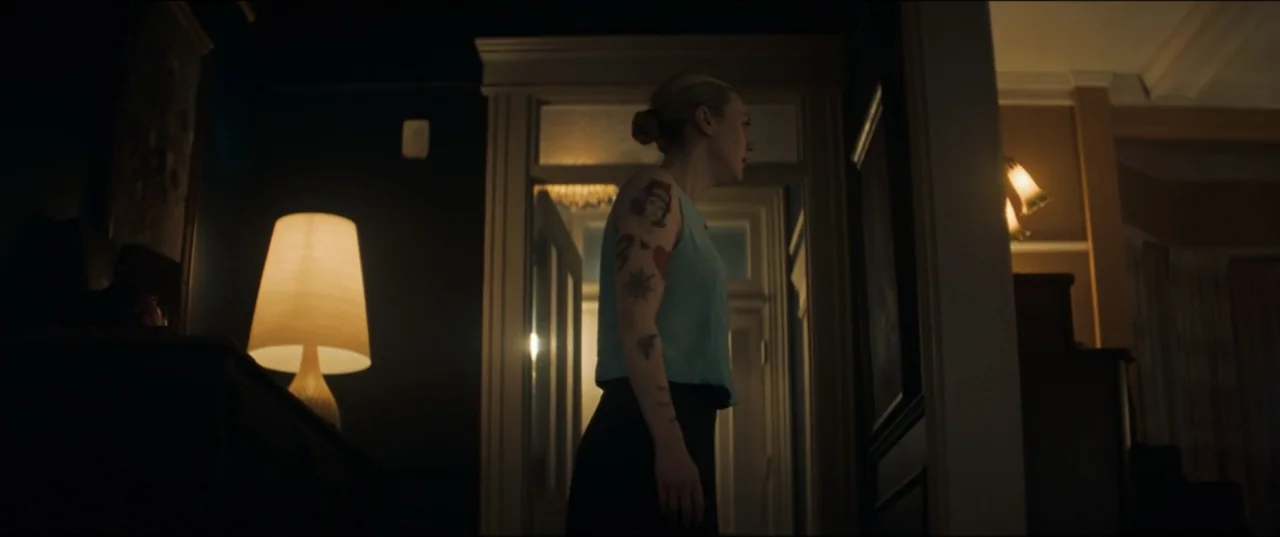 Vicious (2025) movie still: medium — A dark hallway connects rooms, illuminated by a single warm lamp on a sideboard. A young woman with arm tattoos…