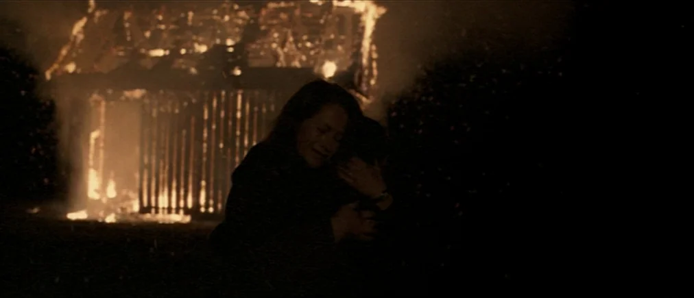 Time Of The Wolf (2003) movie still: medium — A dark outdoor space features a wooden barn engulfed in bright orange flames. A woman hugs a child…