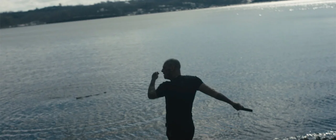 Skin (2009) movie still: fullbody — A man stands on a rocky shore overlooking a large, calm body of water. He holds a small, thin object in his…