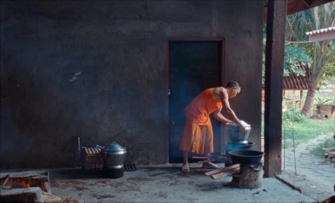 Samsara (2023) movie still: medium — A rustic outdoor cooking space with a concrete wall and an open doorway. A monk in orange robes works at a…