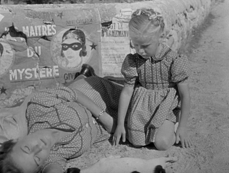 Forbidden Games (1952) movie still: medium — A rural dirt track is lined by a low stone wall covered in posters. A young girl kneels on the sand…