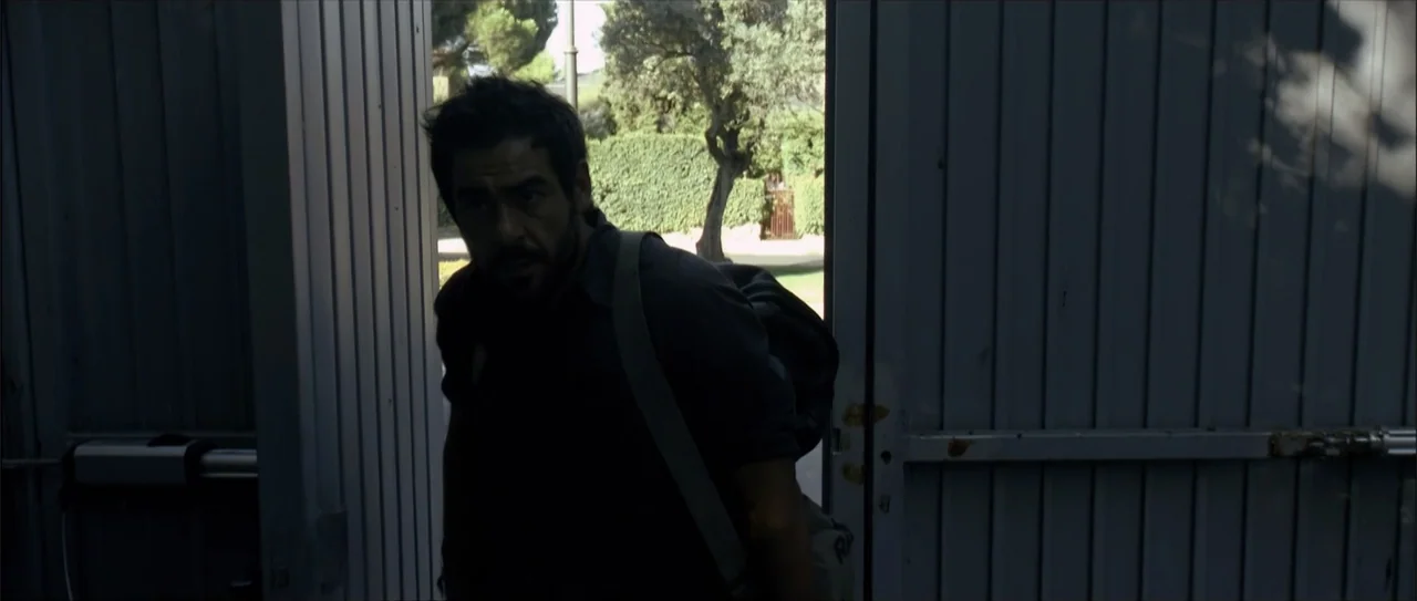 Otro verano (2013) movie still: medium — A man in a dark shirt stands in the shadowed frame of an open gate. He is passing from the dark interior…