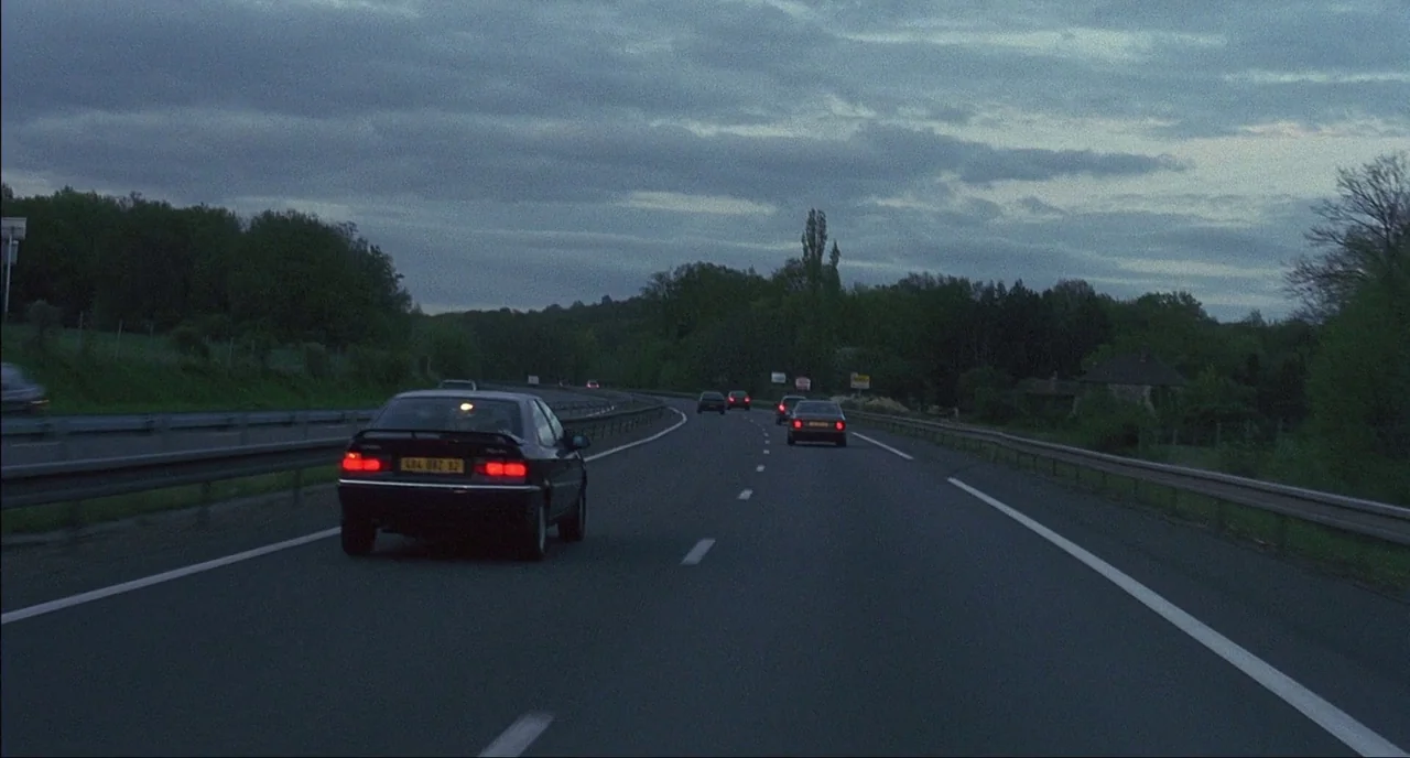 Fat Girl (2001) movie still: wide — A multi-lane asphalt highway curves through a wooded landscape under a cloudy dusk sky. Several passenger cars…