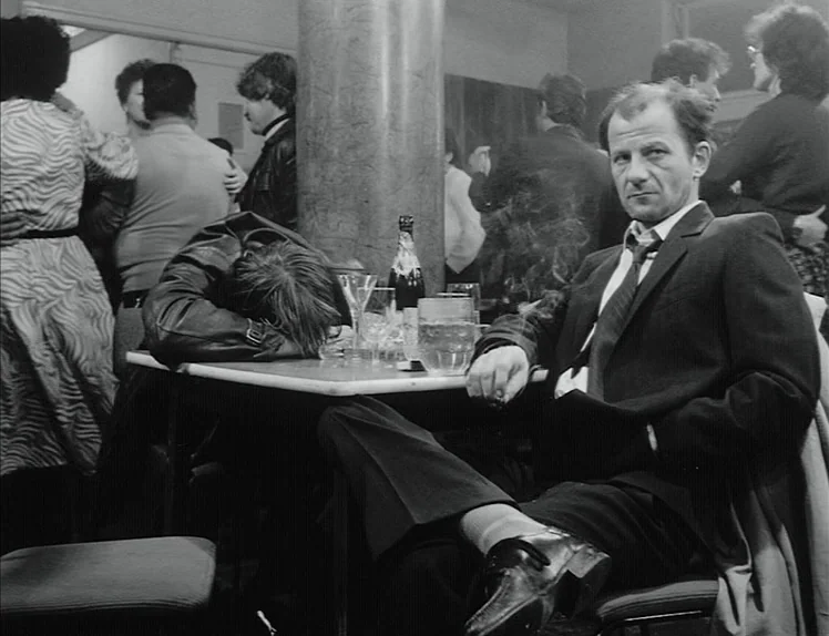 Damnation (1988) movie still: medium — A crowded public social space with people dancing in the soft-focus background area. A man sits at a table in…