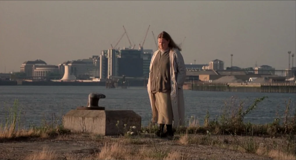 All or Nothing (2002) movie still: fullbody — A woman stands on a sparse dirt riverbank near a concrete mooring bollard. Across the water, a distant…