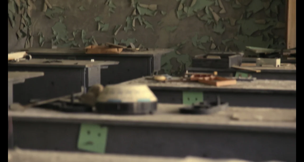 Chernobyl's Café (2016) movie still: medium — An abandoned classroom features rows of dusty, gray desks against a peeling concrete wall. The space…