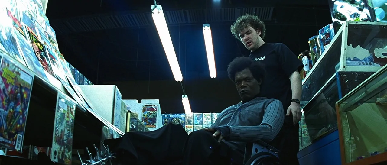 Unbreakable (2000) movie still: medium — A dimly lit comic book store features shelves packed with magazines and glass display cases. A man in a…