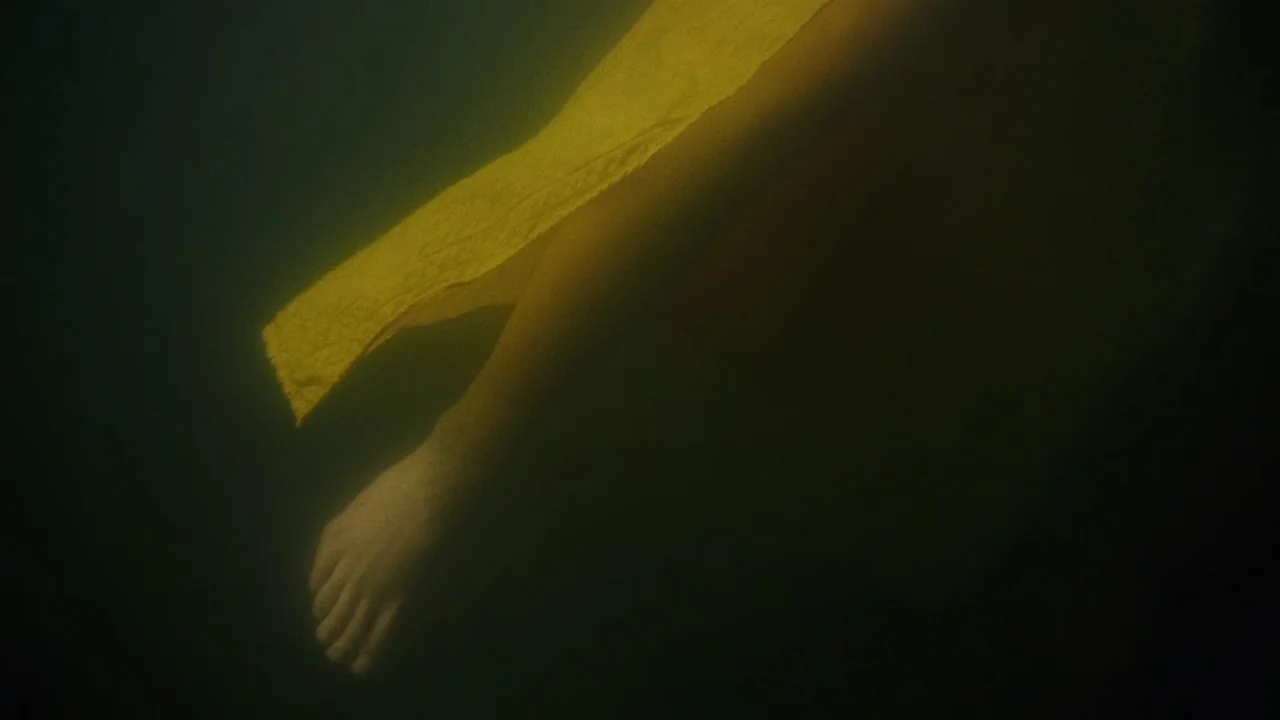 My First Summer (2020) movie still: closeup — A submerged limb floats in dark, murky water with minimal visibility. A bright yellow cloth is wrapped…