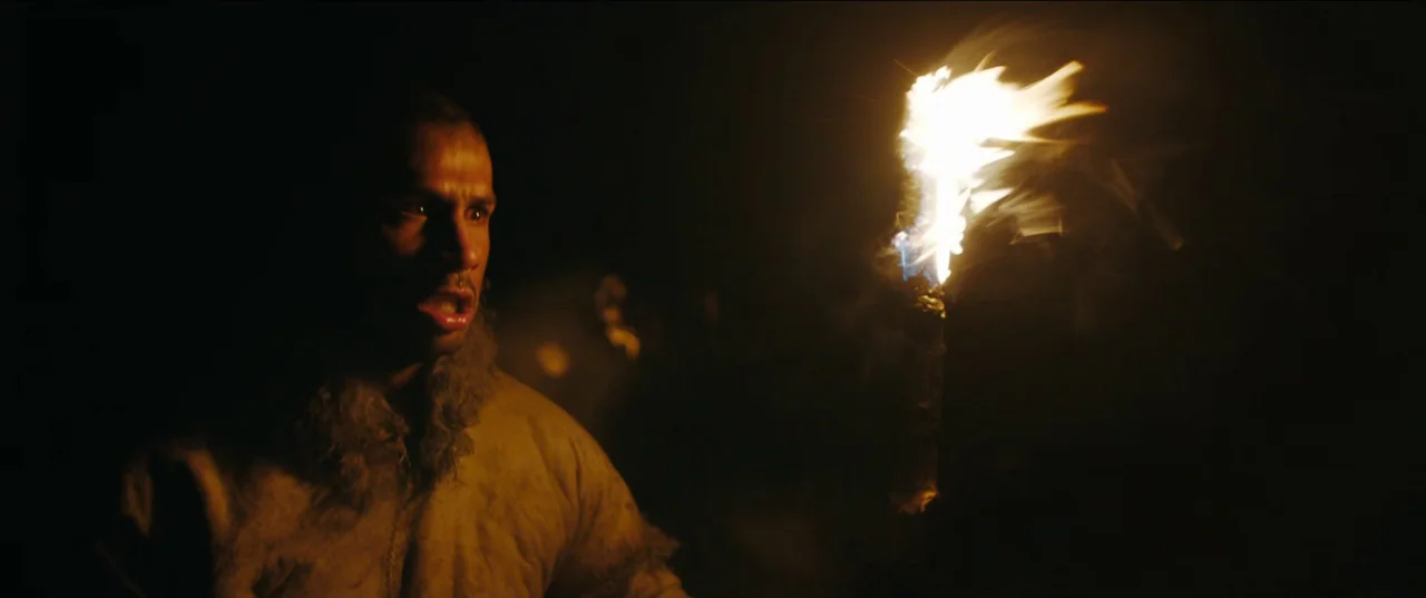 Out of Darkness (2022) movie still: medium — A dark, unidentifiable outdoor space is illuminated only by a nearby hand-held torch. A man with a…