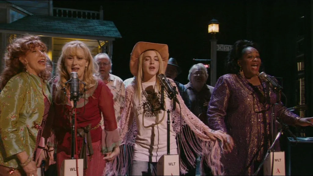 A Prairie Home Companion movie still: medium — A group of singers performs on an outdoor stage at night under a streetlamp. They stand before…