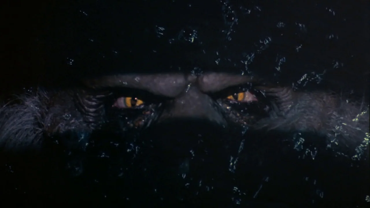 Creepshow (1982) movie still: closeup — A dark, rippling body of water conceals a creature beneath its surface. The creature peers through the black…