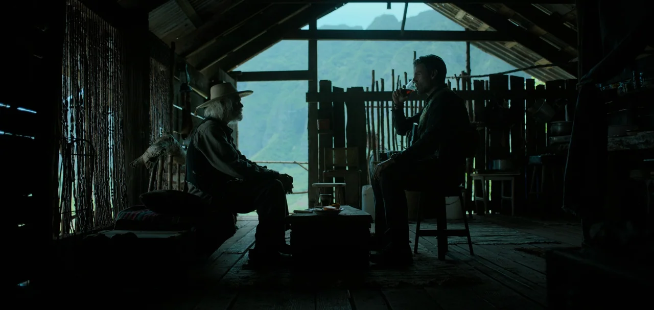 Triple Frontier (2019) movie still: wide — A rustic wooden shack with an open back wall looks out onto a mountain. Two men sit on opposite sides of…