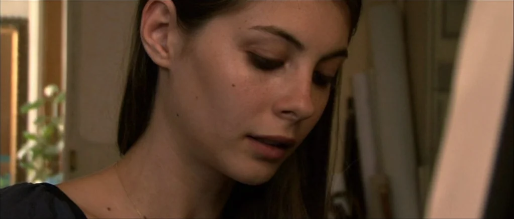 Genova (2008) movie still: closeup — A quiet indoor workspace features stacked canvases and out-of-focus household items. A young woman focuses her…