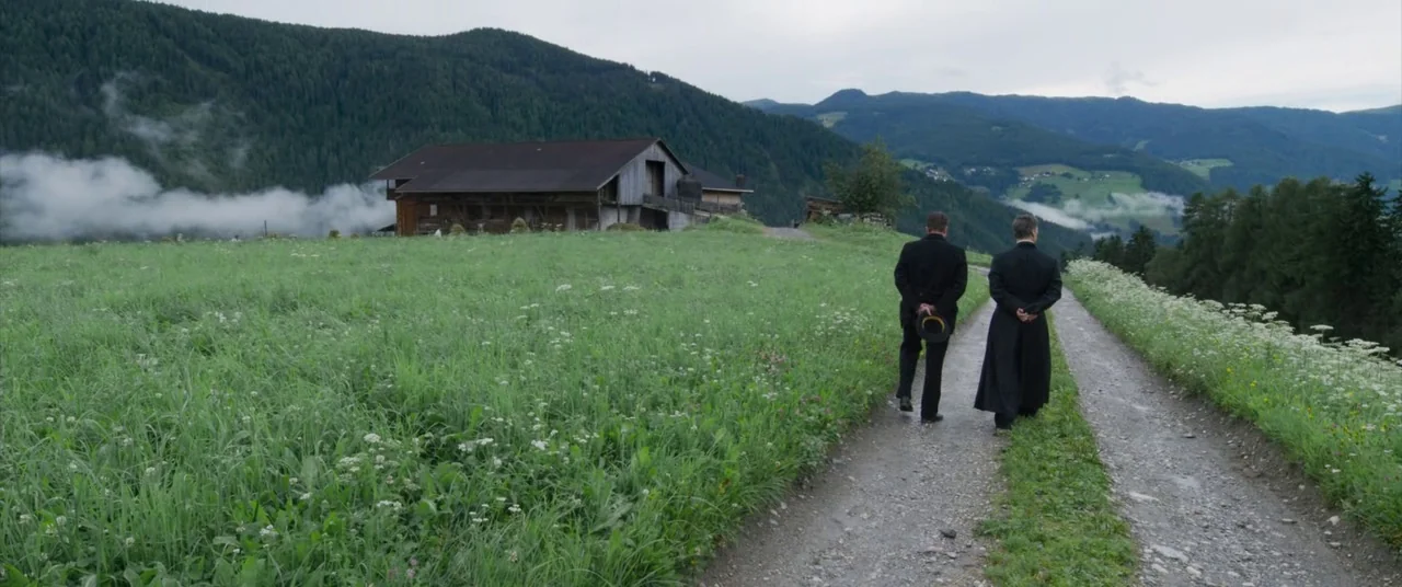 A Hidden Life (2019) movie still: wide — A rural dirt path winds through a verdant meadow toward a wooden mountain structure. Two men in dark formal…