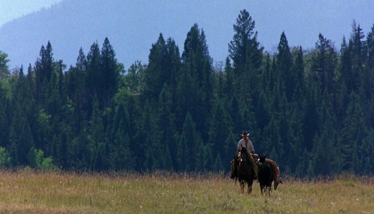 Legends of the Fall (1994) movie still: wide — A man rides a horse across a wide, grassy meadow in front of dense conifers. Tall evergreen trees…