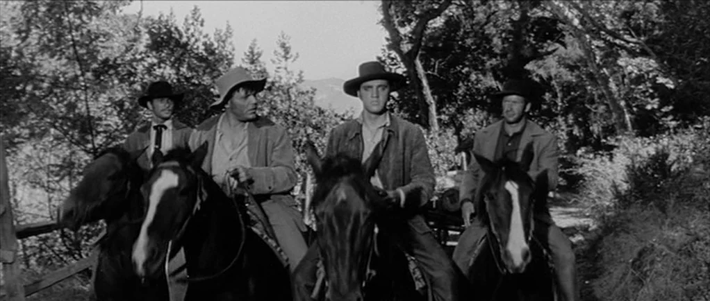 Love Me Tender movie still: medium — Four men ride horses on a dirt trail lined with dense trees and foliage. Each man wears a hat and jacket while…
