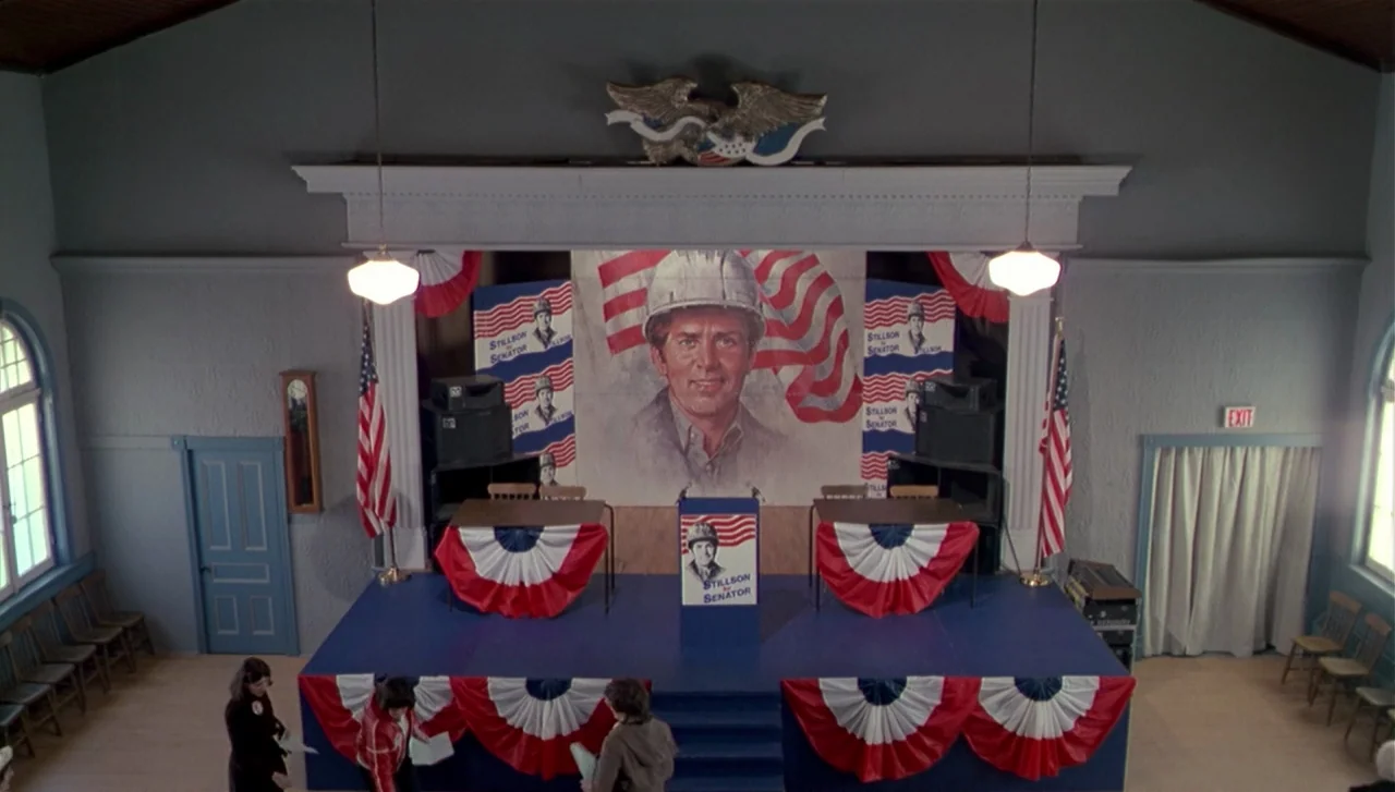 The Dead Zone (1983) movie still: establishing — A high-angle view of a community center hall arranged for a political campaign rally. A blue wooden…
