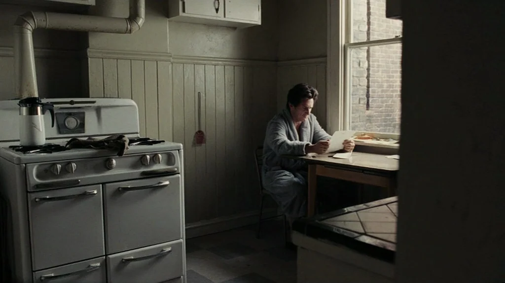 The Assassination of Richard Nixon (2004) movie still: wide — A plain kitchen features wood-paneled walls and a vintage white stove. A man sits at a…