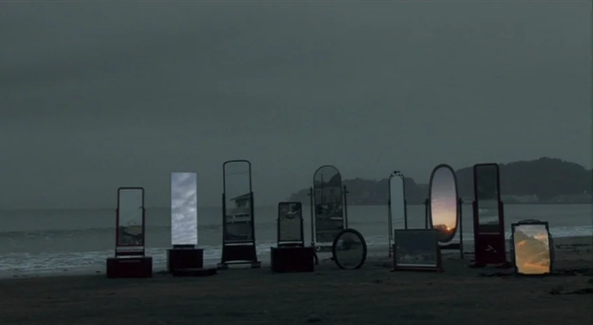 Rampo Noir (2005) movie still: wide — A row of various freestanding mirrors sits on a dark, wet beach at dusk. The mirrors reflect the cloudy sky…