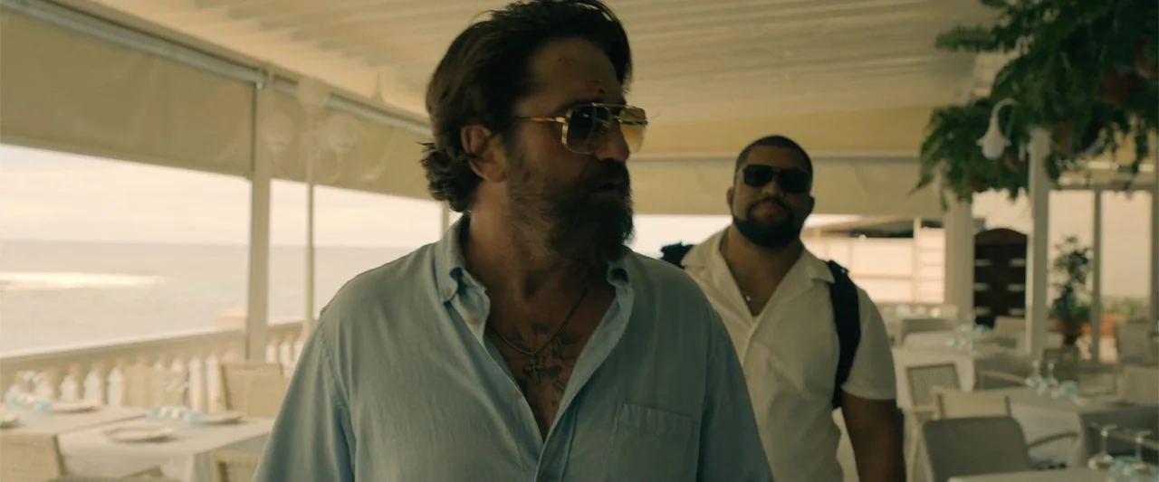 Den of Thieves 2: Pantera (2025) movie still: medium — An open-air restaurant terrace features white tables and a view of the ocean. Two men walk…