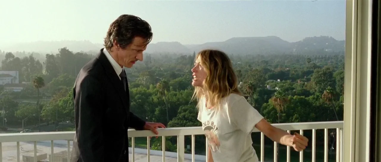Too Late (2015) movie still: medium — An outdoor balcony overlooks a sprawling park with distant hills under hazy sunlight. A woman in a white…