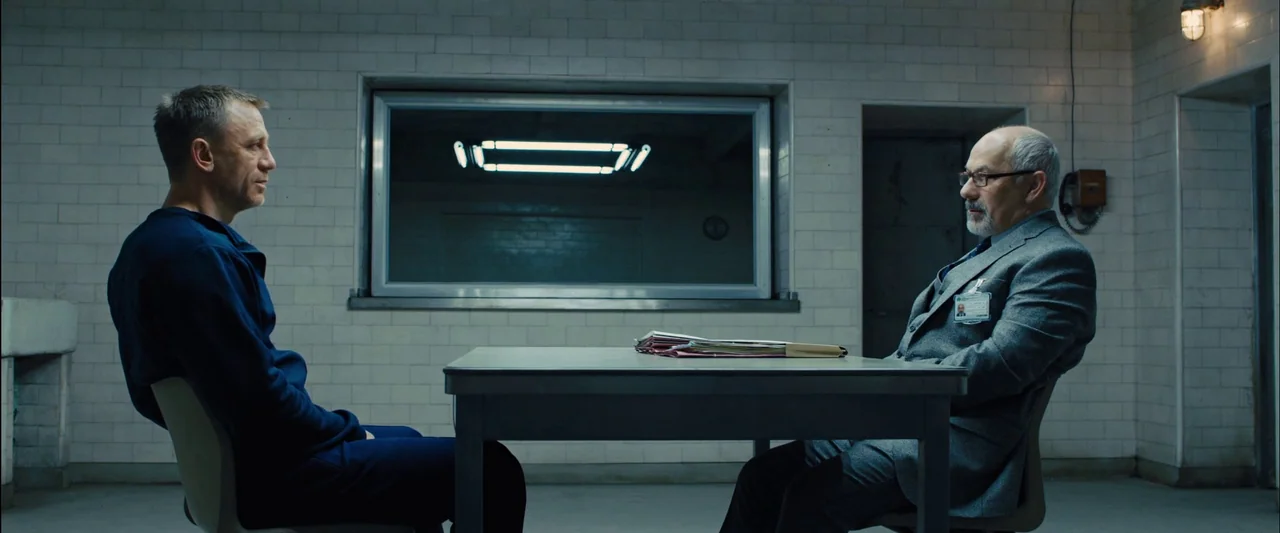Skyfall (2012) movie still: wide — A stark room with floor-to-ceiling white ceramic tiles and a central metal table. Two men sit in chairs facing…