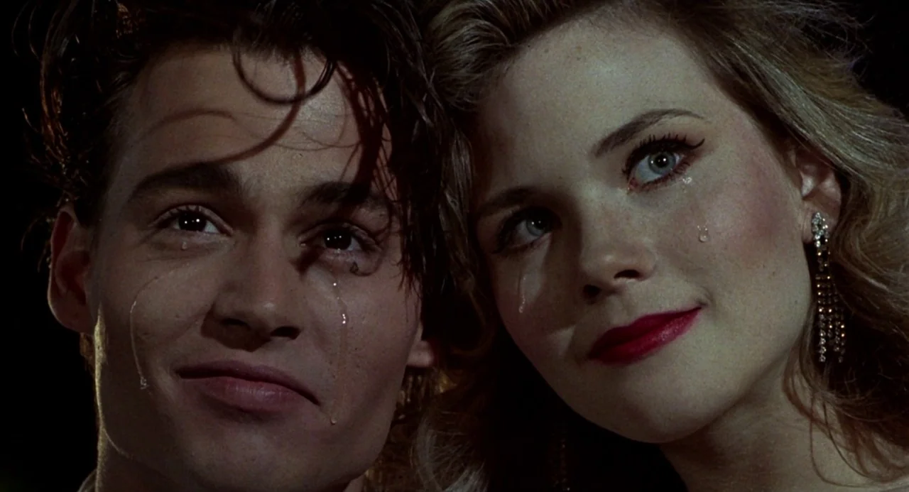 Cry Baby (1990) movie still: closeup — A dark, indistinct indoor space serves as the background for two people side-by-side. The young man and woman…