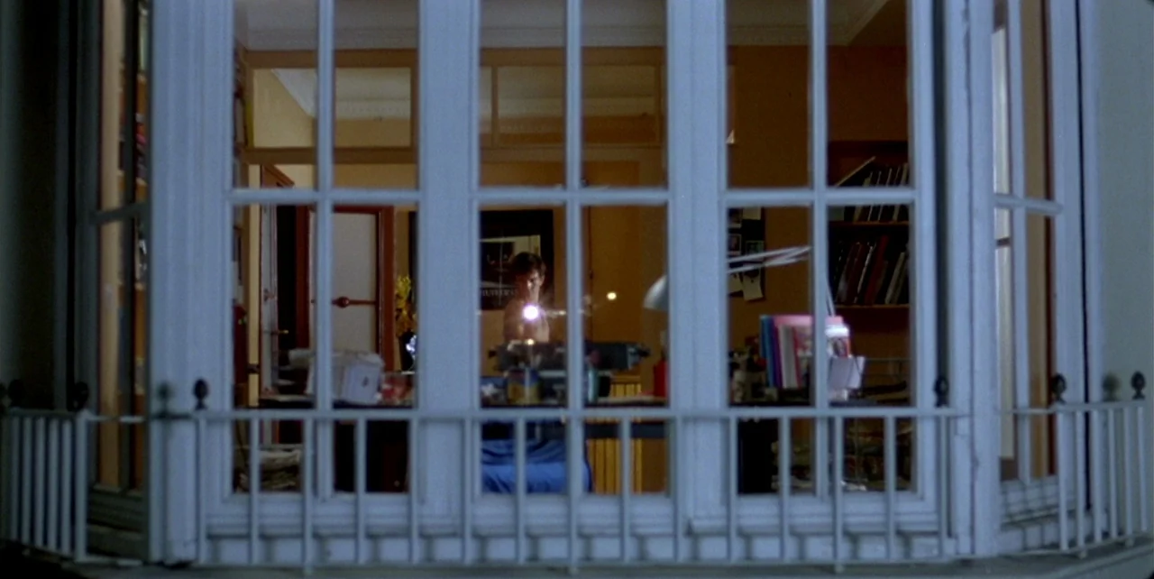 Law of Desire (1987) movie still: wide — A view from outside looking into a building room through a large window. A man sits at a desk while lit by…