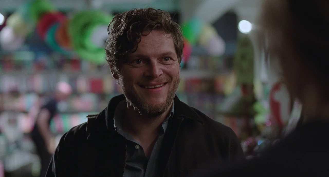 The Worst Person in the World (2021) movie still: medium — A man stands in an aisle of a brightly stocked retail store with blurry merchandise. The…