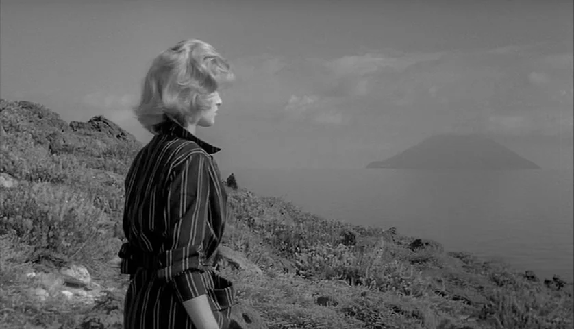 L’Avventura (1960) movie still: medium — A rocky coastal hillside rises above a calm sea under a cloudy sky. A blonde woman stands in the…