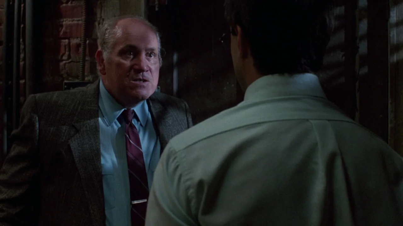 Bride of Reanimator (1989) movie still: medium — A dimly lit industrial hallway features dark exposed brick and vertical iron pipes. A man in a suit…