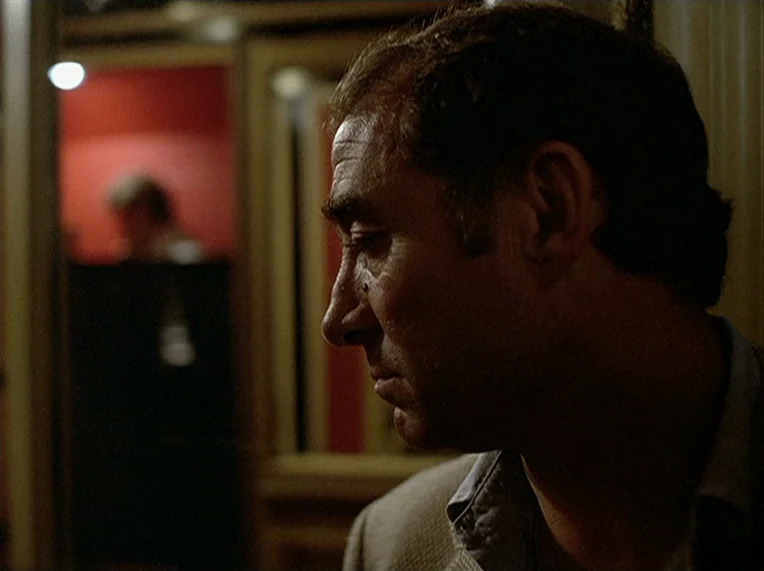 Detective (1985) movie still: closeup — A dimly lit interior space with a red-walled background visible in the distance. A man is positioned in the…