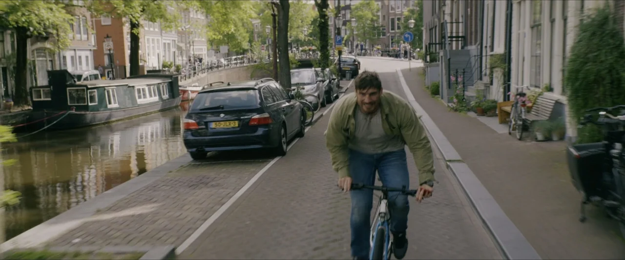 Black Lotus (2023) movie still: medium — A paved path runs alongside a canal in a European-style city with parked cars. A man rides a bicycle…