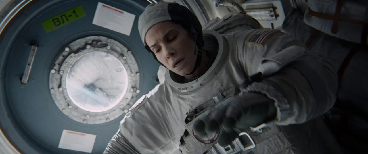 Gravity (2013) movie still: medium — A cramped spacecraft interior features a circular porthole with a label marked VL-1. A female astronaut in a…