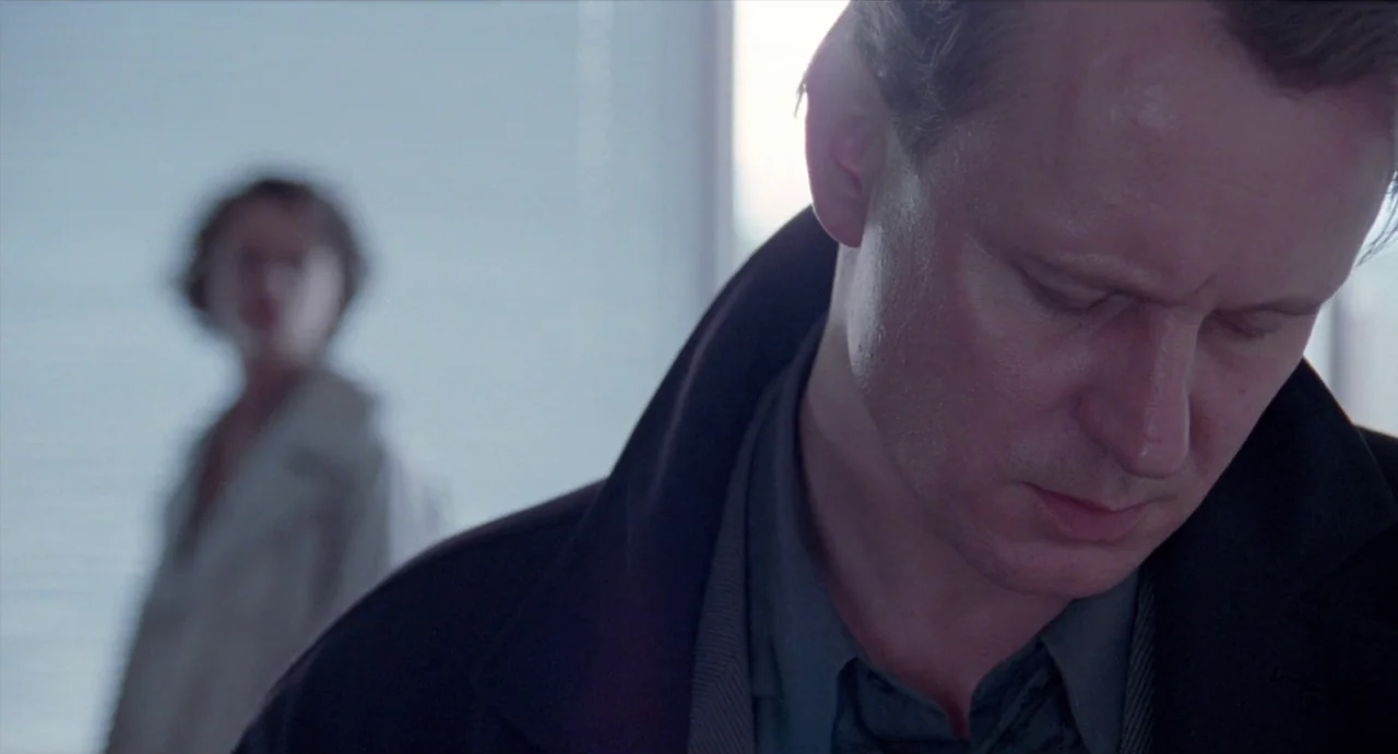 Insomnia (1997) movie still: closeup — An interior space with a bright, diffused window background and minimalist architecture. A middle-aged man in…