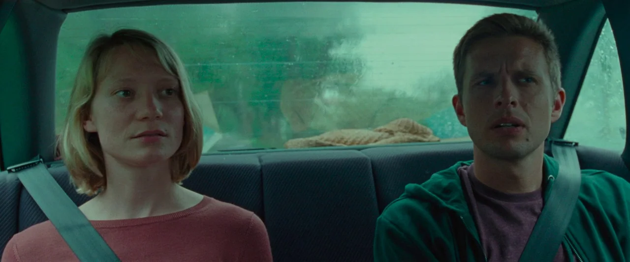 Bergman Island (2021) movie still: medium — A view from the backseat of a car during an overcast day. A woman and a man sit belted in side by side.